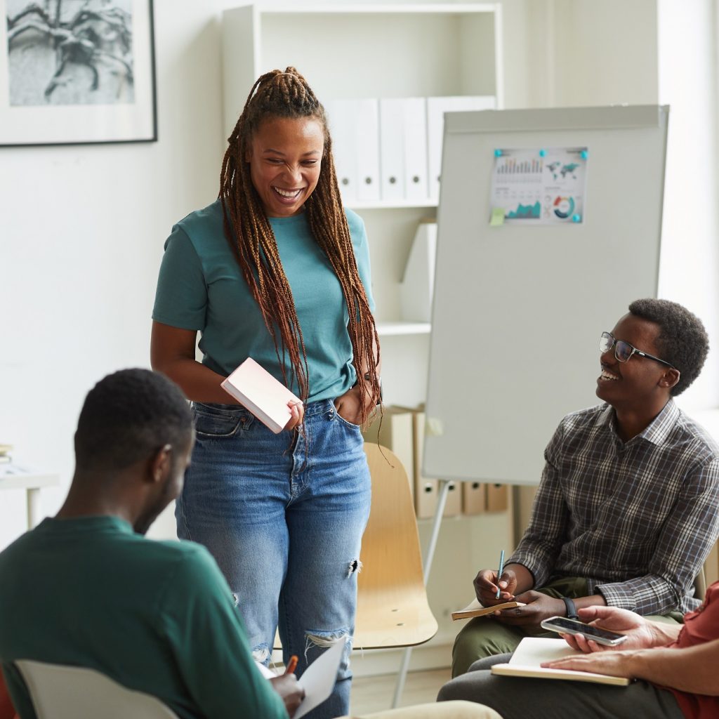 Multi-ethnic group of people sitting in circle while discussing business project in office, focus on smiling African-American woman talking to colleagues, copy space